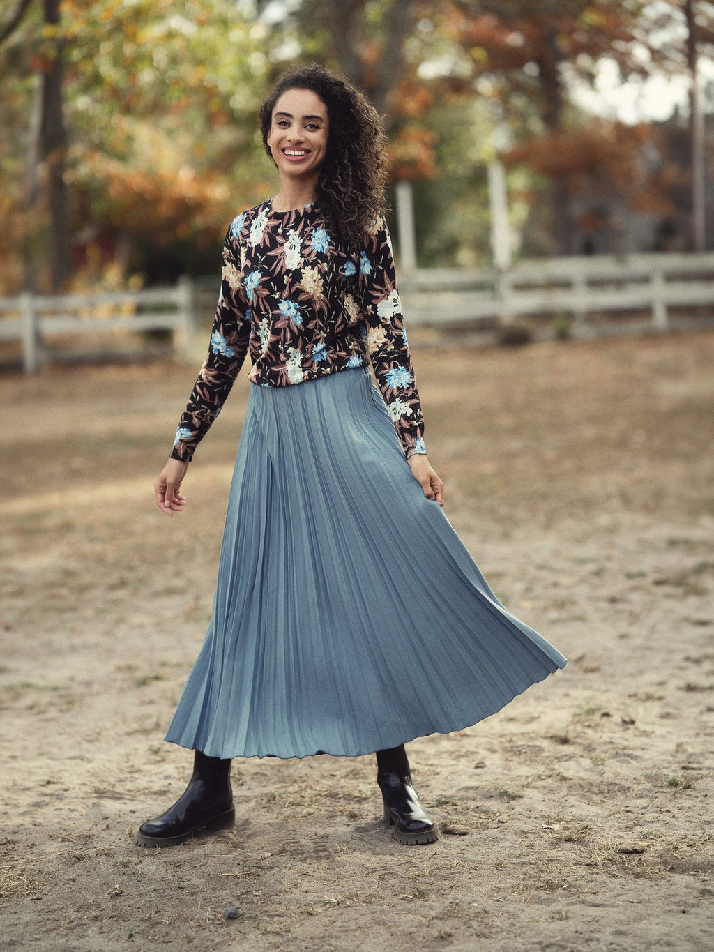 Woman wearing a floral sweater and blue pleated skirt standing in an outdoor setting with trees and a fence.