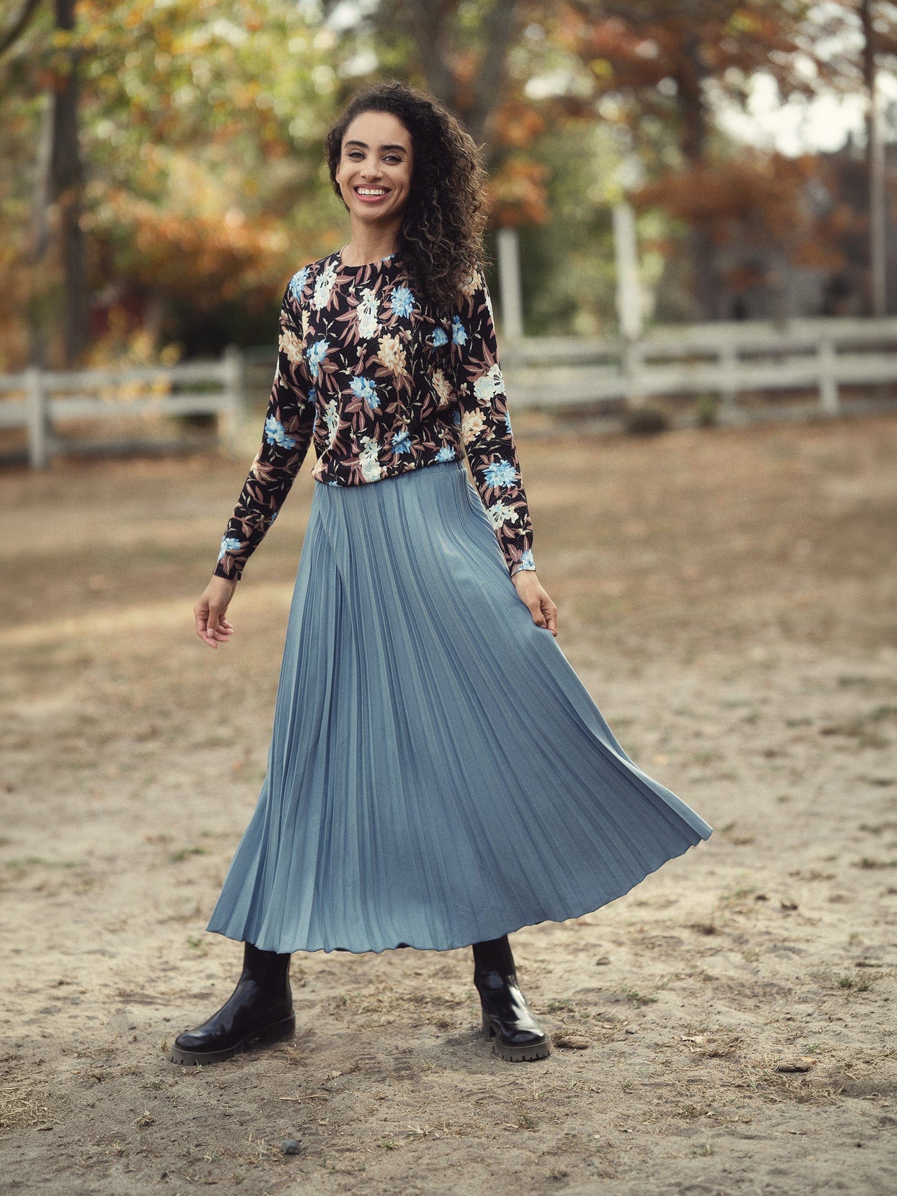 Woman wearing a floral sweater and blue pleated skirt standing in an outdoor setting with trees and a fence.