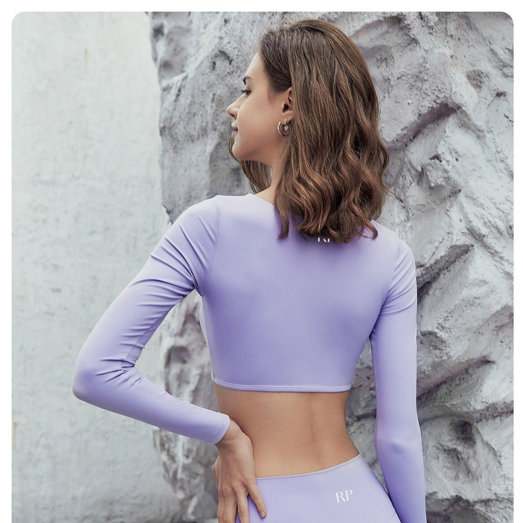 Woman wearing a purple long-sleeve crop top and leggings against a textured wall.