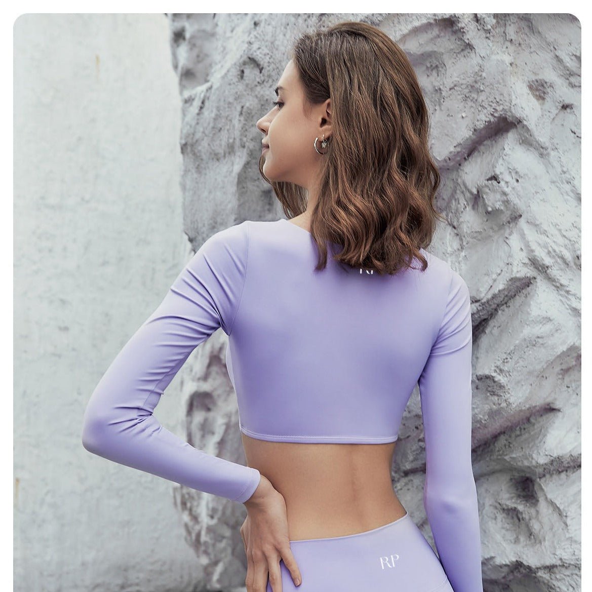 Woman wearing a purple long-sleeve crop top and leggings against a textured wall.
